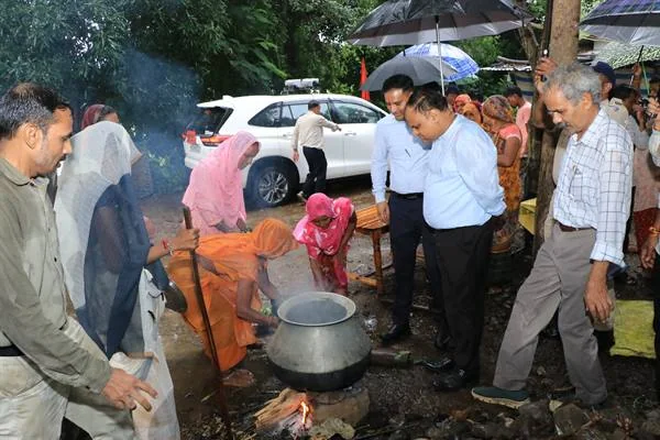 बारिश के बीच पेड़ों के नीचे चूल्हे पर रखे बड़े बर्तन के पास खड़े लोग