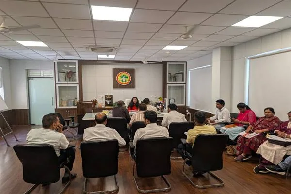 कार्यालय के बैठक कक्ष में कुर्सियों पर बैठे अधिकारी और सामने मेज पर बैठी महिला अधिकारी