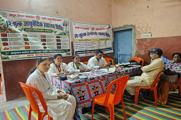 एक हॉल में मेज़ के पीछे बैठीं सफ़ेद एप्रन पहने तीन महिला चिकित्सक और एक पुरुष कर्मचारी, मेज़ पर दवाइयों की शीशियाँ रखी हैं; सामने नारंगी कुर्सियों पर दो बुज़ुर्ग बैठे हैं और दीवार पर आयुष विभाग मध्यप्रदेश के निःशुल्क आयुर्वेदिक और होम्योपैथी स्वास्थ्य शिविर के बैनर लगे हैं।